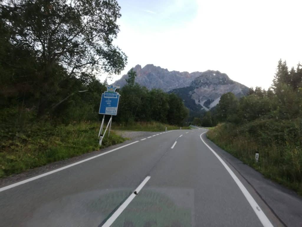 Eine kurvige Bergstraße führt an einem blauen Schild vorbei auf ein mächtiges Bergmassiv zu