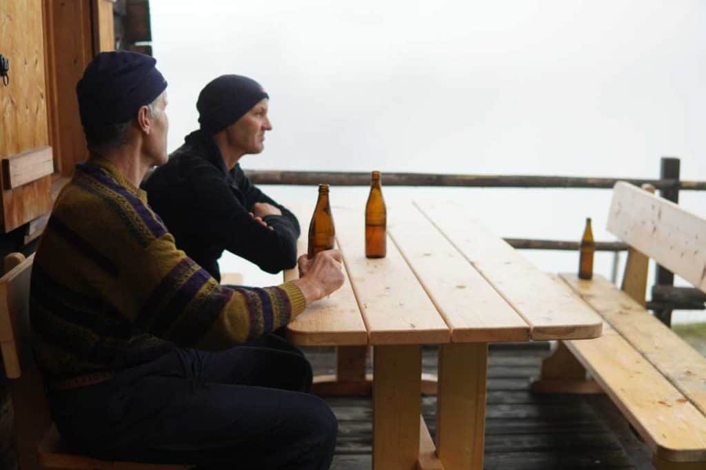 Zwei Männer mit Hauben sitzen mit Bierflaschen an einem Holztisch im Nebel