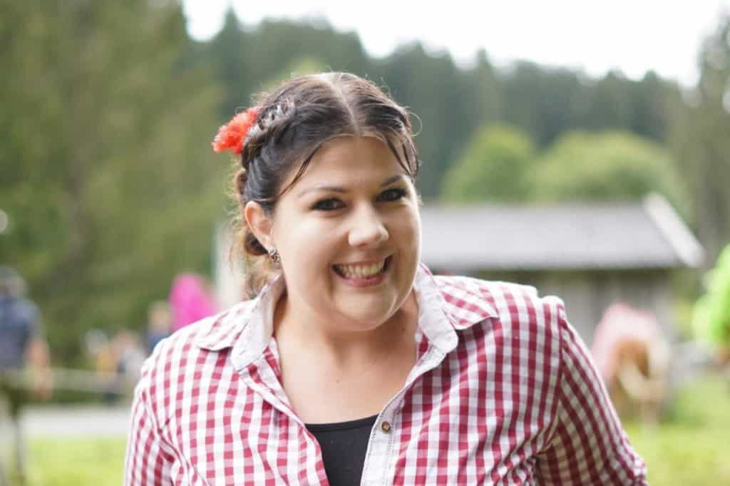 Eine lächelnde Frau im rot-weiß karierten Hemd blickt vor Bäumen und einer Hütte direkt in die Kamera