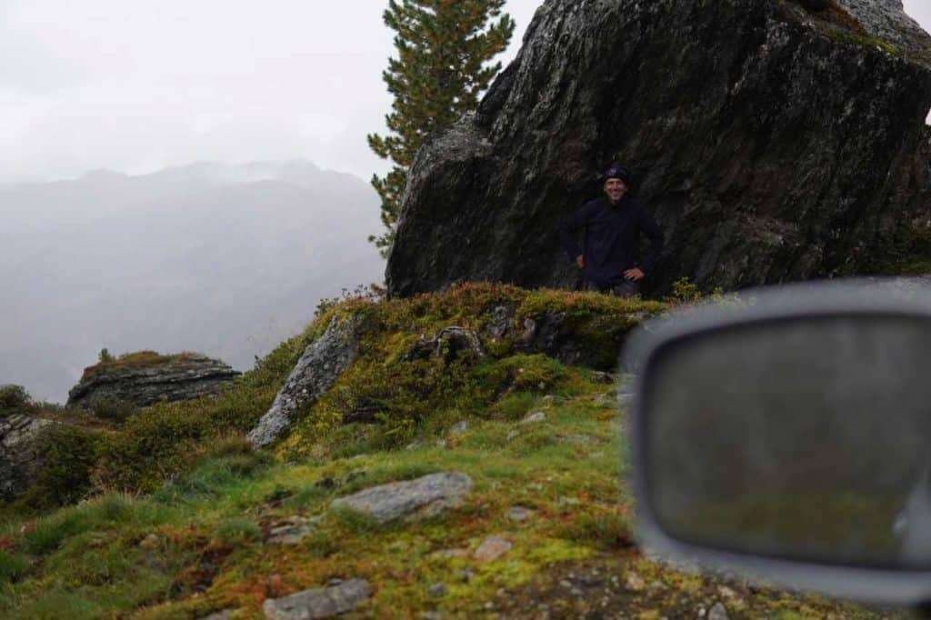 Blick am Seitenspiegel vorbei auf eine Person vor einem Felsen in einer nebeligen Almlandschaft