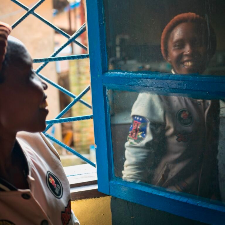 Zwei lachende Frauen an einem blauen Fenstergitter, eine davon spiegelt sich in der Scheibe